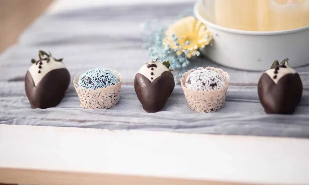 Detailaufnahme von süßen Hochzeitshäppchen: Schoko-Erdbeeren im Smoking-Look und Pralinen mit blauen Streuseln auf einem Tuch dekoriert