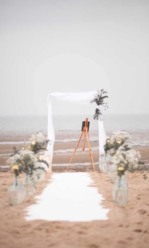 Stimmungsvolles Hochzeits-Setup am Strand mit weißem Traubogen, Blumenarrangements in Glasvasen und Blick auf das Meer